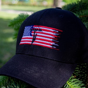 Puede incluir: Una gorra de béisbol negra con un diseño de bandera estadounidense desgastado que presenta un poste de línea eléctrica y cables. La bandera es roja, blanca y azul.