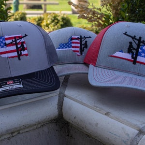 May include: Three gray and red trucker hats with a silhouette of a person working on a power line with an American flag design. The hats are on a gray stone surface.