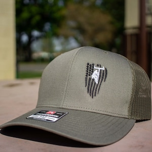May include: A green and brown trucker hat with a black and white embroidered American flag design featuring a silhouette of a lineman. The flag is tattered and the lineman is climbing a utility pole.