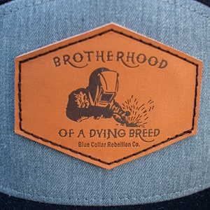 以下が含まれることがあります： 「Brotherhood of a Dying Breed」というテキストと溶接工のシルエットが描かれた茶色の革パッチ。「Blue Collar Rebellion Co.」というテキストがメインテキストの下にある。