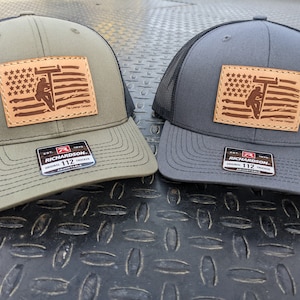 May include: Two trucker hats with a black mesh back. One hat is green with a brown leather patch featuring an American flag with a lineman climbing a pole. The other hat is grey with the same patch. Both hats have a Richardson 112 label.