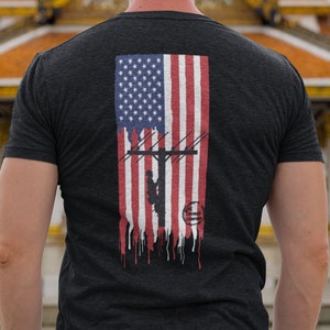 Puede incluir: Camiseta gris oscuro con un diseño de la bandera estadounidense y un liniero escalando un poste de electricidad. El diseño de la bandera tiene un efecto desgastado y goteante. Un logotipo circular en la parte inferior.