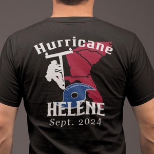 Op de afbeelding: Zwart T-shirt met een witte afbeelding van een elektricien die aan een stroomlijn werkt met een rode en witte omtrek van de staat Florida. De tekst "Hurricane Helene Sept. 2024" is onder de afbeelding gedrukt.