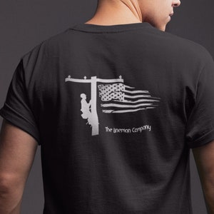 Puede incluir: Camiseta negra con una silueta blanca de un lineman escalando un poste de electricidad con una bandera estadounidense detrás de él. El texto "The Lineman Company" está impreso debajo de la imagen.