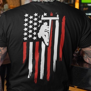 Puede incluir: Camiseta negra con una silueta blanca de un electricista escalando un poste de electricidad frente a un diseño de bandera estadounidense.