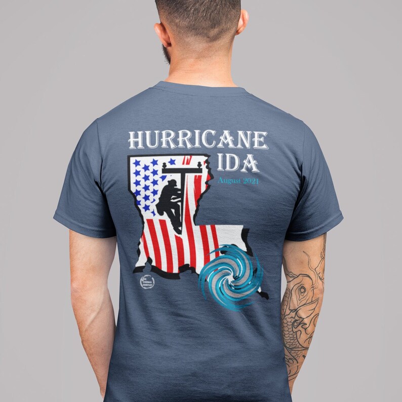 Pu&ograve; includere: Una maglietta blu con una grafica che commemora l'uragano Ida. Il design include il testo "HURRICANE IDA August 2021" sopra una raffigurazione del contorno dello stato della Louisiana, la bandiera americana e un uragano stilizzato.