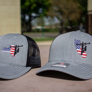 May include: Two gray and black trucker hats with an American flag and a silhouette of a lineman on a power pole. The hats are embroidered with the state outlines of New York and California.