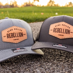 Op de afbeelding: Twee trucker caps met zwarte mesh achterpanelen. De caps zijn grijs en zwart. Beide caps hebben een bruin leren patch met de tekst "Blue Collar Rebellion Company" erop.