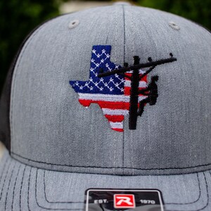 Può includere: Un cappellino da baseball grigio mélange con retro in rete nera. La parte anteriore presenta un ricamo dello stato del Texas con il motivo della bandiera americana, sovrapposto alla sagoma nera di un palo della luce e di un operaio.