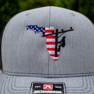 May include: A gray and black trucker hat with an embroidered design of a power line pole and a lineman, with the American flag in the background. The hat has a black mesh back and a gray front with the text "EST. 1970 RICHARDSON" on the bottom.