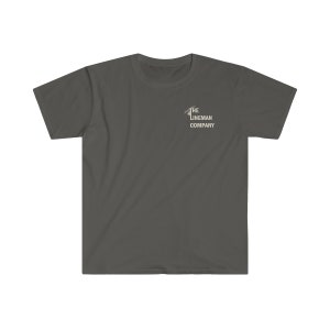 Op de afbeelding: Een donkergrijs T-shirt met een witte afbeelding van een monteur die een elektriciteitsmast beklimt. De afbeelding bevat de tekst "The Lineman Company".