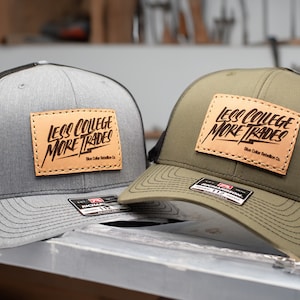 Op de afbeelding: Twee trucker caps met zwarte mesh achterpanelen. De ene pet is grijs met een bruin leren patch waarop "Less College More Trades" staat en de andere pet is groen met dezelfde patch.