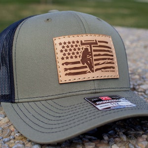 Può includere: Un cappello da camionista verde e nero con una toppa in pelle marrone che presenta la silhouette di un elettricista che lavora su una linea elettrica davanti a un design di bandiera americana. La toppa ha il testo "The Lineman Company" nell'angolo in basso a destra.