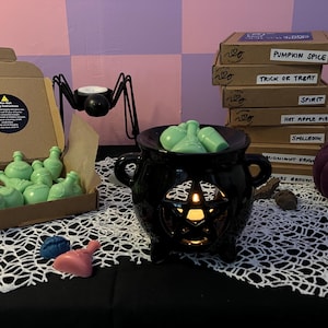 May include: A black cauldron wax melt warmer with a pentagram design, surrounded by green potion wax melts. A stack of brown boxes with the text "Pumpkin Spice", "Trick or Treat", "Spirit", "Hot Apple Pie", "Spellbook", "Midnight Potion", and "Witches Broom" are visible in the background.