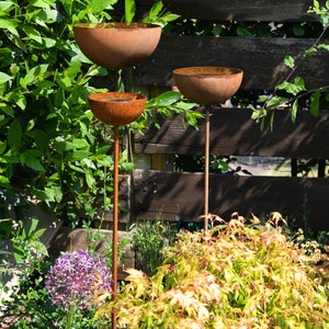 May include: Three rusty metal bowls on tall metal stems, arranged in a tiered formation. The bowls are positioned in a garden setting with green foliage and a wooden fence in the background.