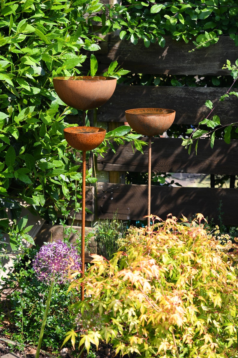 May include: Three rusty metal bowls on tall metal stems, arranged in a tiered formation. The bowls are positioned in a garden setting with green foliage and a wooden fence in the background.