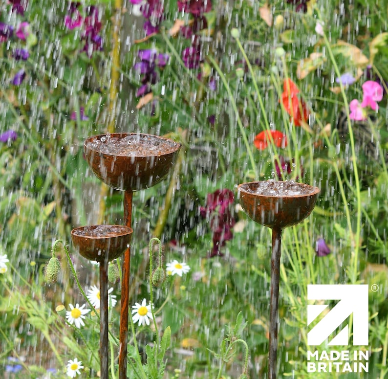 May include: Three copper-colored metal bird feeders on tall metal stems in a garden setting. The feeders are shaped like bowls and are catching raindrops. The background is a blurred image of green foliage and colorful flowers.