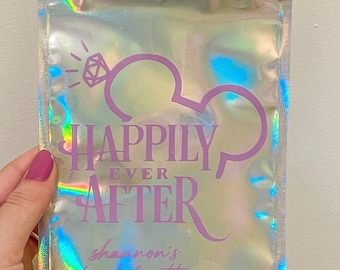 May include: A rectangular holographic pouch with a resealable top. The front features purple text that reads "Happily Ever After" with a diamond icon and mouse ear silhouette, plus the text "shannon's bachelorette" in a cursive script font.