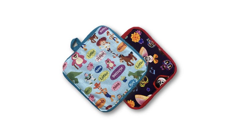 May include: Two square pot holders. One is light blue with cartoon characters and the other is dark blue with skulls and flowers. Both have a loop for hanging and are trimmed in a contrasting color.