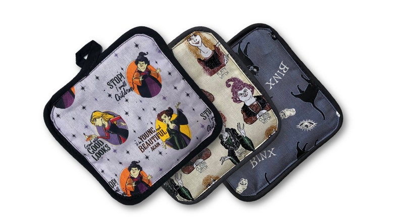 Puede incluir: Tres agarraderas de olla de tela gris y negro con tem&aacute;tica de Halloween. Las agarraderas presentan im&aacute;genes de brujas y un gato negro con el texto "Stop I will Children", "The Young and Beautiful Again", y "Binx".