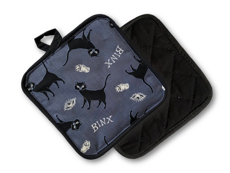 Puede incluir: Un par de agarraderas para ollas. Una es negra con textura acolchada. La otra es azul con un patr&oacute;n de gatos negros, ojos y la palabra "BINX".
