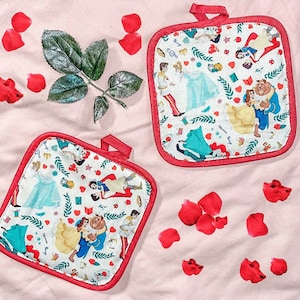 May include: Two square, white pot holders with red trim and hanging loops. The pot holders feature a colorful pattern of Disney characters, including Belle and the Beast, and other fairytale elements. Red rose petals and a green leaf are scattered around the pot holders on a pink surface.