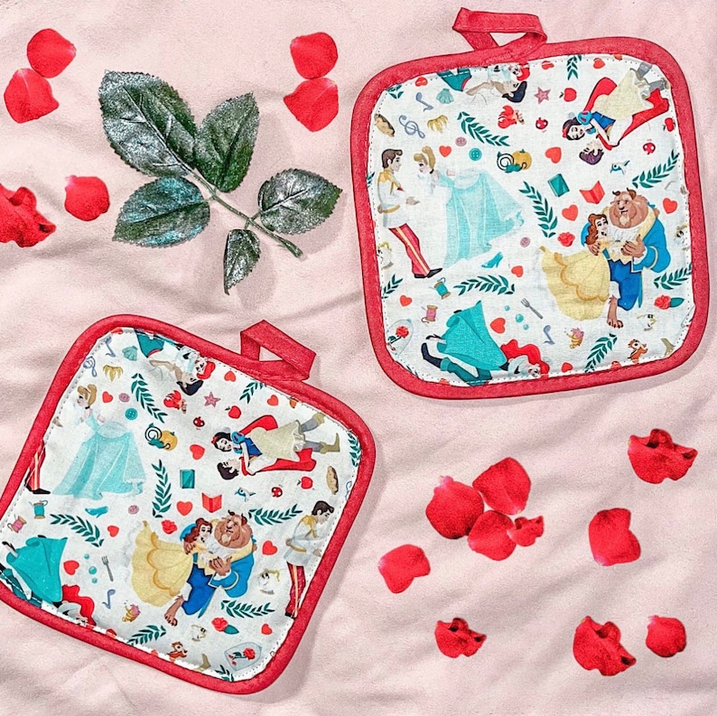 May include: Two square, white pot holders with red trim and hanging loops. The pot holders feature a colorful pattern of Disney characters, including Belle and the Beast, and other fairytale elements. Red rose petals and a green leaf are scattered around the pot holders on a pink surface.