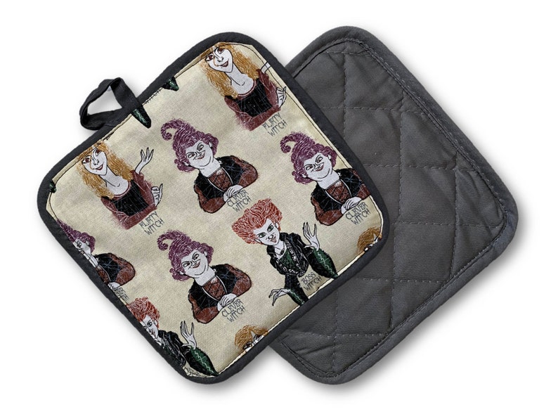 Puede incluir: Un par de guantes para horno con una parte posterior acolchada negra y una parte delantera de tela beige con un patr&oacute;n repetido de brujas de dibujos animados con el texto "Clever Witch" y "Pretty Witch".