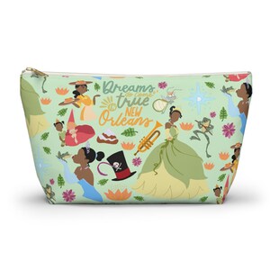May include: A green zippered pouch with a repeating pattern of Tiana and other characters from the Disney movie The Princess and the Frog. The text "Dreams do come true in New Orleans" is printed on the pouch.