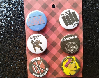 Puede incluir: Conjunto de seis botones redondos con diferentes bandas de punk rock. Los botones son blancos con texto y gráficos en negro y rojo. Las bandas que aparecen son: Black Flag, The Ramones, Blondie, Dead Kennedys, Operation Ivy y Adolescents.