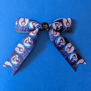 May include: A hair bow made of blue and black ribbon featuring a repeating graphic print of a cartoon character with blue hair against a moonlit background. The center of the bow is secured with a small, round, black four-hole button.