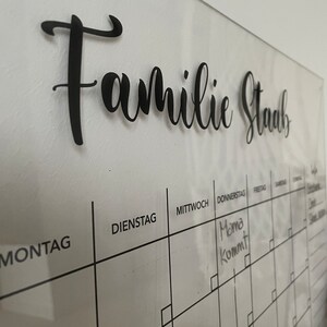 May include: A white dry-erase calendar with a black grid and the words "Familie Staub" at the top. The days of the week are labeled in German: Montag, Dienstag, Mittwoch, Donnerstag, Freitag, Samstag, Sonntag. The word "Mama kommt" is written in black marker on the Thursday square.