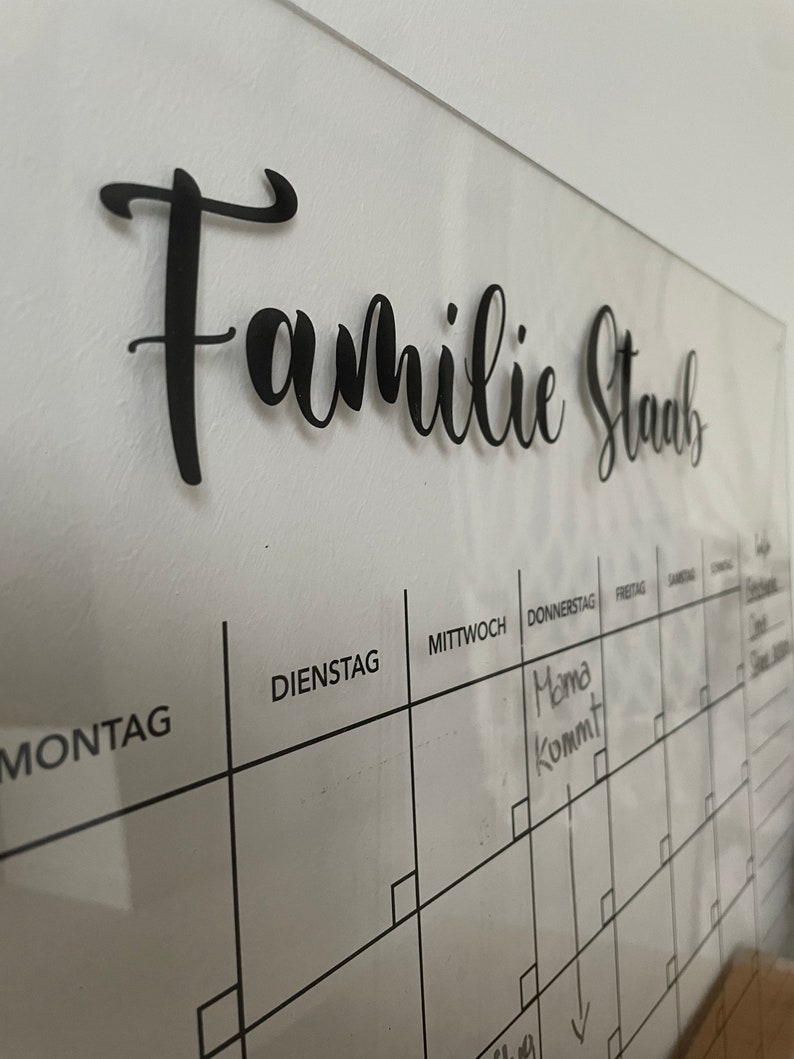 May include: A white dry-erase calendar with a black grid and the words "Familie Staub" at the top. The days of the week are labeled in German: Montag, Dienstag, Mittwoch, Donnerstag, Freitag, Samstag, Sonntag. The word "Mama kommt" is written in black marker on the Thursday square.