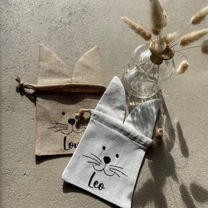 Puede incluir: Dos peque&ntilde;as bolsas de regalo de tela con orejas de conejo. Las bolsas est&aacute;n hechas de yute y algod&oacute;n blanco. La bolsa de yute tiene la palabra "Love" escrita en ella. La bolsa blanca tiene la palabra "Leo" escrita en ella.