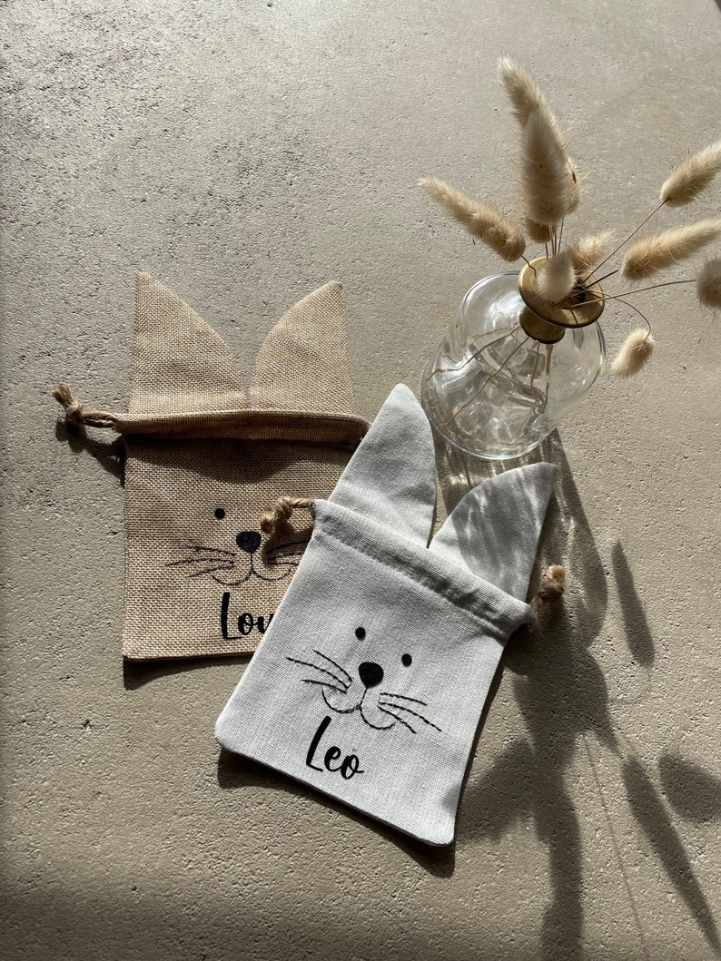 Puede incluir: Dos peque&ntilde;as bolsas de regalo de tela con orejas de conejo. Las bolsas est&aacute;n hechas de yute y algod&oacute;n blanco. La bolsa de yute tiene la palabra "Love" escrita en ella. La bolsa blanca tiene la palabra "Leo" escrita en ella.