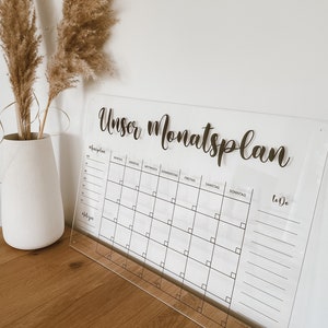 May include: A clear acrylic monthly planner with the text "Unser Monatsplan" at the top. The planner has a grid for each day of the week, with spaces for notes and a to-do list.