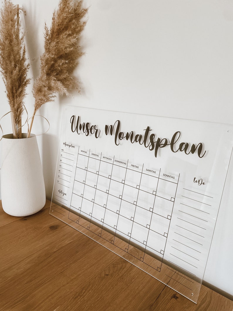 May include: A clear acrylic monthly planner with the text "Unser Monatsplan" at the top. The planner has a grid for each day of the week, with spaces for notes and a to-do list.