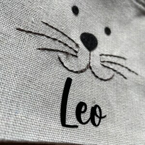 Puede incluir: Bolsa de lona blanca con una cara de gato bordada en negro y el nombre "Leo" escrito debajo.