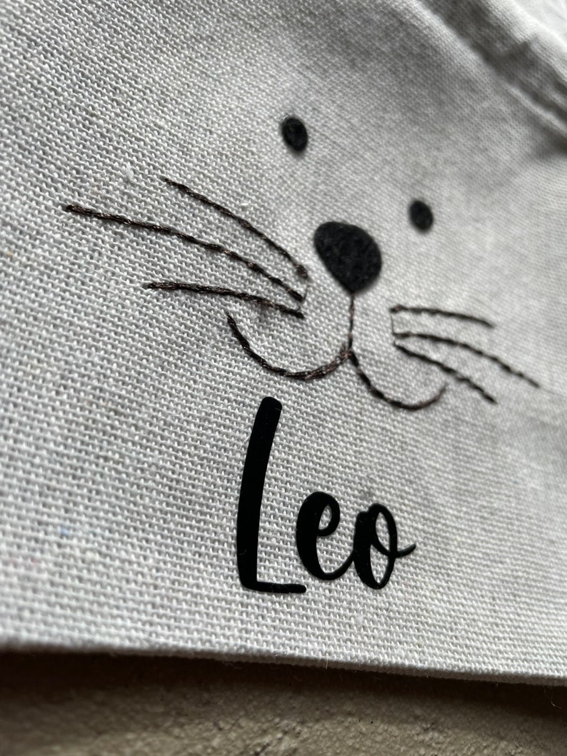 Puede incluir: Bolsa de lona blanca con una cara de gato bordada en negro y el nombre "Leo" escrito debajo.
