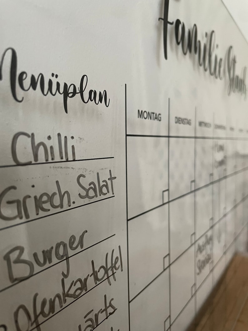 May include: A white dry-erase board with a black marker listing a weekly meal plan. The meal plan includes "Chilli", "Greek Salad", "Burger", "Potato", and "Arts". The board also has a calendar with the days of the week labeled in German: "Montag" (Monday), "Dienstag" (Tuesday), and "Mittwoch" (Wednesday).