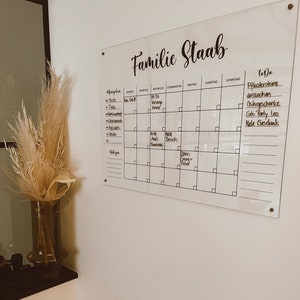 May include: A white acrylic dry-erase board with a black calendar grid and to-do list. The board has the text "Familie Staab" written at the top. The calendar grid has the days of the week labeled in German. The to-do list includes items such as "Pflastersteine aussuchen" and "Ostergeschenke".