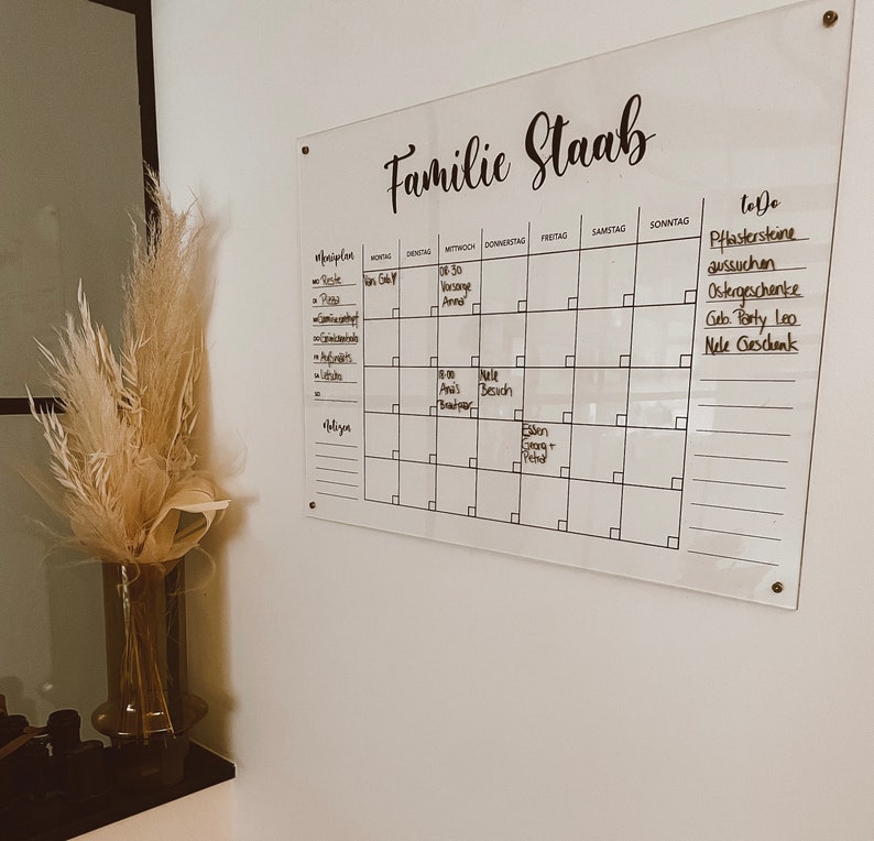 May include: A white acrylic dry-erase board with a black calendar grid and to-do list. The board has the text "Familie Staab" written at the top. The calendar grid has the days of the week labeled in German. The to-do list includes items such as "Pflastersteine aussuchen" and "Ostergeschenke".