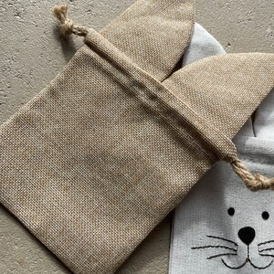 Puede incluir: Una bolsa de regalo de arpillera con cierre de cord&oacute;n y un dise&ntilde;o de cara de gato. La bolsa es blanca con rasgos de gato negros y el nombre "Leo" impreso debajo de la cara.
