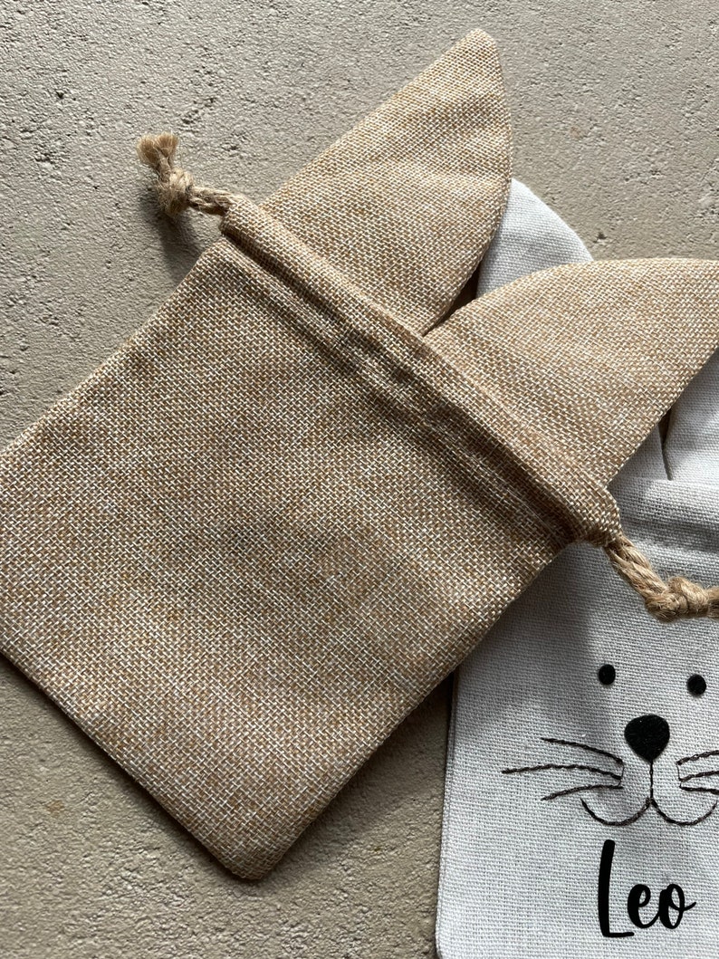 Puede incluir: Una bolsa de regalo de arpillera con cierre de cord&oacute;n y un dise&ntilde;o de cara de gato. La bolsa es blanca con rasgos de gato negros y el nombre "Leo" impreso debajo de la cara.