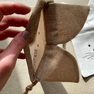 Puede incluir: Una peque&ntilde;a bolsa de arpillera marr&oacute;n con cierre de cord&oacute;n. La bolsa est&aacute; abierta y el interior es visible. La bolsa est&aacute; hecha de fibras naturales y tiene un aspecto r&uacute;stico.