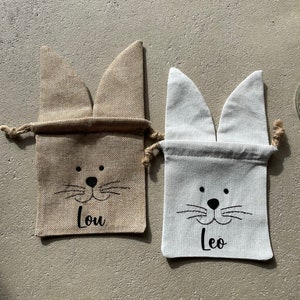 Puede incluir: Dos peque&ntilde;as bolsas de regalo de arpillera con orejas de conejo. La bolsa de la izquierda es marr&oacute;n y tiene el nombre "Lou" impreso en ella. La bolsa de la derecha es blanca y tiene el nombre "Leo" impreso en ella.