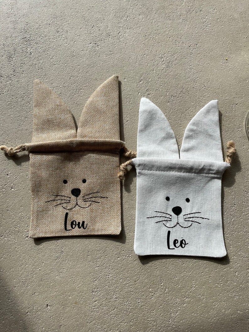 Puede incluir: Dos peque&ntilde;as bolsas de regalo de arpillera con orejas de conejo. La bolsa de la izquierda es marr&oacute;n y tiene el nombre "Lou" impreso en ella. La bolsa de la derecha es blanca y tiene el nombre "Leo" impreso en ella.