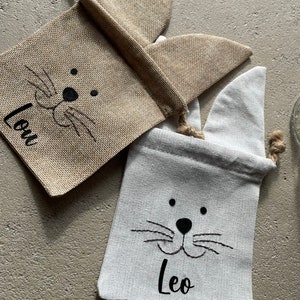 Puede incluir: Dos bolsas de regalo de conejito de arpillera con caras de conejito bordadas en negro y los nombres "Leo" y "Lou" en ellas.