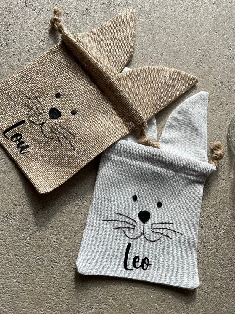 Puede incluir: Dos bolsas de regalo de conejito de arpillera con caras de conejito bordadas en negro y los nombres "Leo" y "Lou" en ellas.