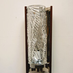 K&ouml;nnte beinhalten: Eine Wandleuchte im Stil der Mid-Century Modern mit einem klaren Glasschirm mit geometrischem Muster. Die Leuchte hat einen Rahmen aus Holz und Metall.